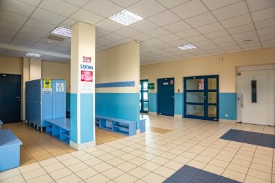 Szkolna szatnia z niebieskimi szafkami, ławkami, wieszakami i wejściem do windy
