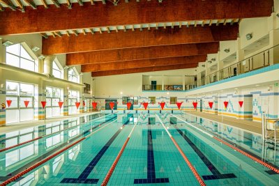 Kryty basen sportowy z torami pływackimi, czerwonymi chorągiewkami i drewnianymi belkami pod sufitem