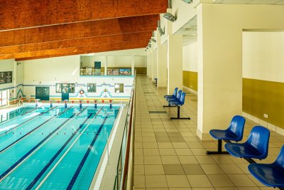 Kryta pływalnia z torami basenowymi i niebieskimi krzesełkami na galerii widokowej